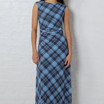 A woman stands against a white brick wall wearing a sleeveless, knee-length blue plaid dress with a gathered waist and black flats.