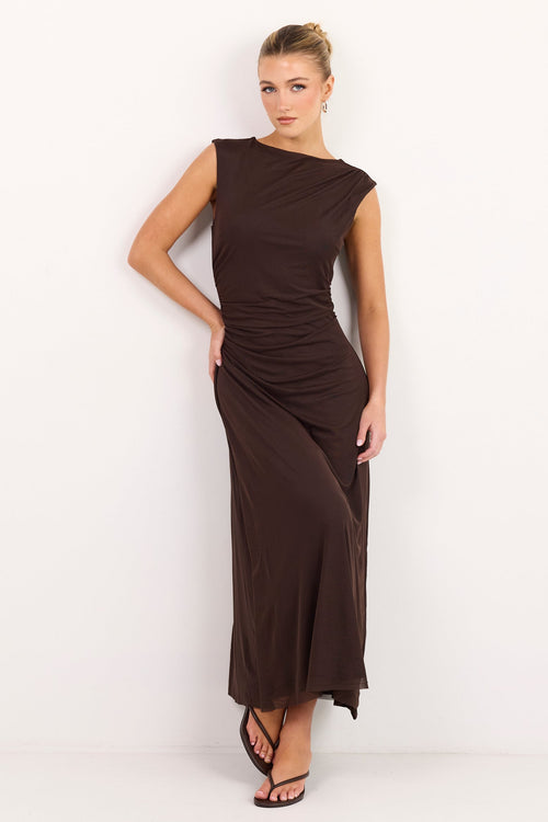 A woman modeling a sleeveless, dark brown maxi dress with an asymmetrical draped neckline and gathered waist, standing against a plain white studio background.