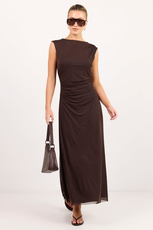 A woman modeling a sleeveless, floor-length dark brown dress with gathered ruching at the waist, styled with dark sunglasses, a matching handbag, and dark sandals against a plain white studio background.