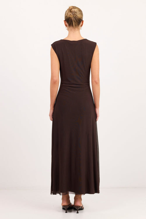 A woman viewed from behind wearing a sleeveless dark brown maxi dress with a gathered waist, standing against a plain white studio background.