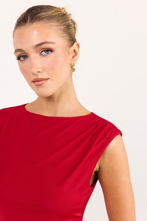 A woman wearing a bright red sleeveless top with a draped neckline, photographed against a clean off-white studio background.