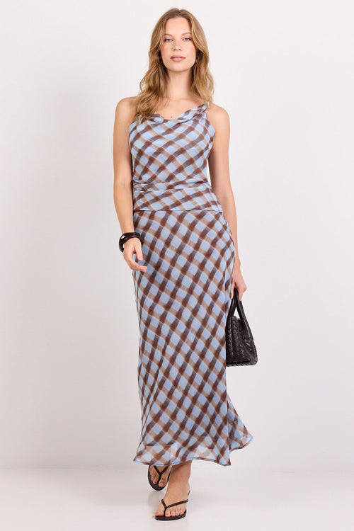 A woman modeling a sleeveless, floor-length halter dress with a brown and gray checkered pattern, holding a small black handbag against a plain white studio background.