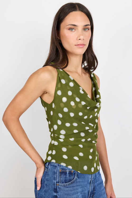A woman modeling a sleeveless olive green wrap-style top with a white polka dot pattern, paired with blue jeans, against a plain light gray studio background.