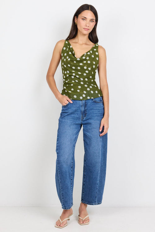 A woman modeling a sleeveless olive green top with a white polka-dot pattern, paired with blue jeans and beige sandals, standing against a plain white studio background.