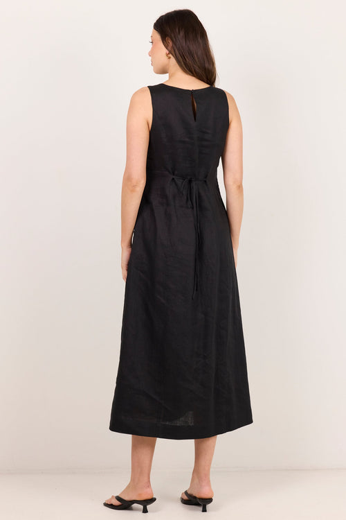 A woman wearing a sleeveless black midi dress with a tie waist, shown from the back against a plain off-white studio background.