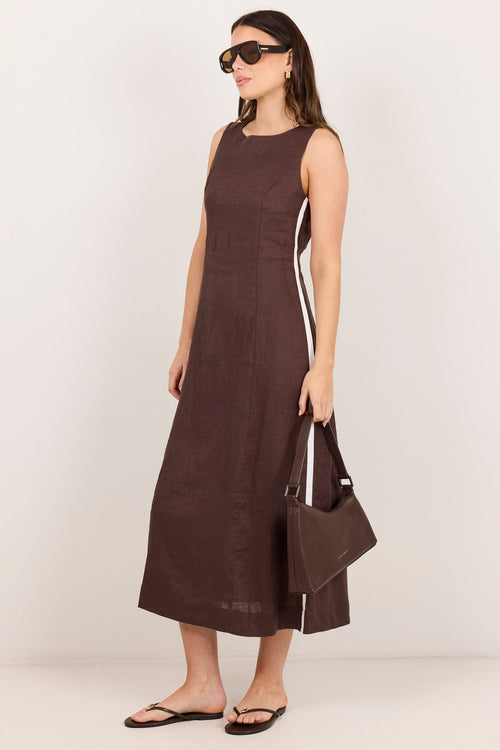 A woman modeling a sleeveless dark brown midi dress with white side stripes, holding a matching brown handbag and wearing sunglasses and flip-flops against a plain off-white studio background.