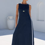 A woman wearing a sleeveless navy maxi dress with a white side stripe and a gold button detail, standing in a sunny outdoor courtyard with white walls and green trees in the background.