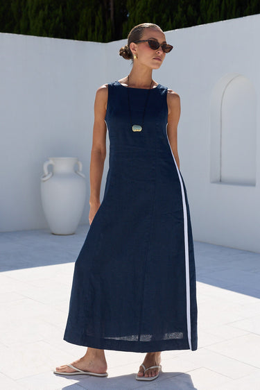 A woman wearing a sleeveless navy maxi dress with a white side stripe and a gold button detail, standing in a sunny outdoor courtyard with white walls and green trees in the background.