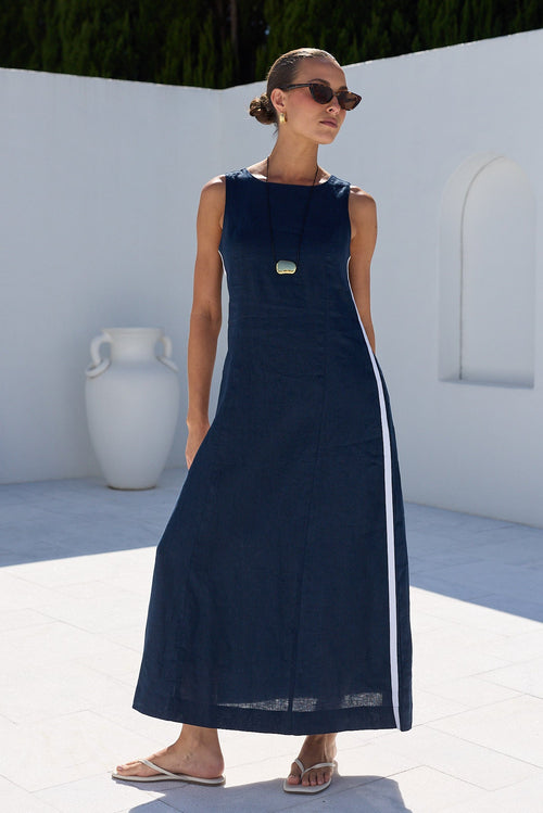 A woman wearing a sleeveless navy maxi dress with a white side stripe and a gold button detail, standing in a sunny outdoor courtyard with white walls and green trees in the background.