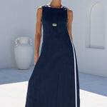 A woman modeling a sleeveless navy blue maxi dress with a white side stripe, standing in a bright, modern outdoor setting with white walls and dark green trees.