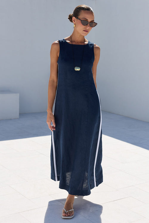 A woman modeling a sleeveless navy maxi dress with white side stripes, standing in a minimalist white outdoor setting.