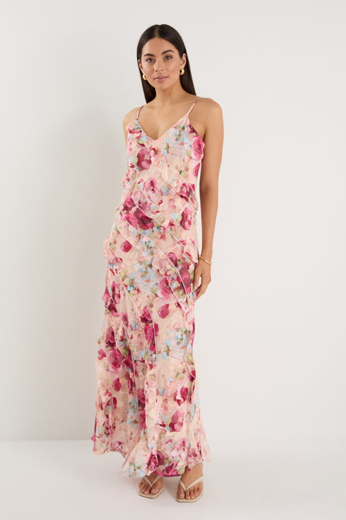 Woman wearing a strappy floral bias-cut maxi dress in pink and pastel tones with cascading ruffle layers, styled with neutral sandals.