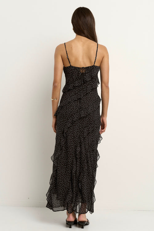 Woman wearing a black bias-cut polka dot dress with delicate straps and cascading ruffles, standing against a neutral wall.