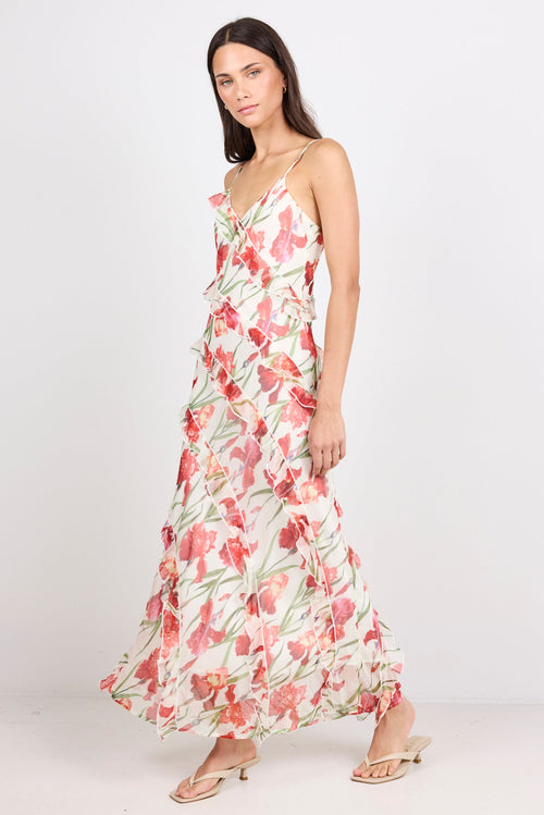 A woman modeling a sleeveless maxi dress with a ruffled bodice and skirt, featuring a pink and red floral print on a white background, paired with beige flat sandals against a white studio backdrop.