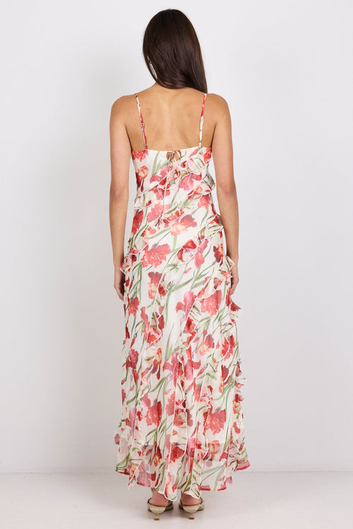 A woman viewed from behind wearing a sleeveless maxi dress with thin straps and a red, pink, and green floral print on a light background, standing against a plain white studio backdrop.