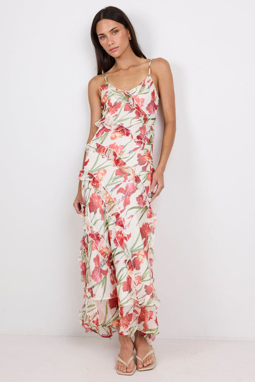 A woman modeling a sleeveless maxi dress with thin straps and a red, pink, and green floral print on a light background, standing against a plain white studio backdrop.