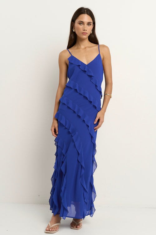 Woman wearing a bright electric blue bias-cut maxi dress with delicate straps and cascading ruffle details, standing against a neutral background.