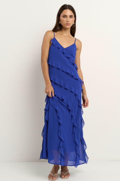 Woman wearing a bright electric blue bias-cut maxi dress with delicate straps and cascading ruffle details, standing against a neutral background.