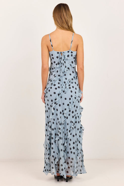A woman viewed from the back wearing a light blue sleeveless maxi dress with black polka dots and thin shoulder straps, standing against a plain off-white studio background.