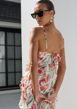 A woman wearing a white spaghetti-strap maxi dress with a red and green tropical floral print, styled with black sunglasses, gold earrings, and a cream crescent handbag, posing outdoors against a white architectural background.