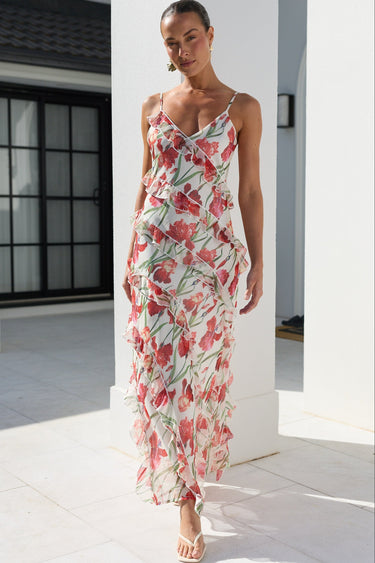 A woman models a sleeveless, ruffled maxi dress with a red and pink floral print on a white background, walking in a bright, modern white outdoor corridor.