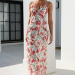 A woman models a sleeveless, ruffled maxi dress with a red and pink floral print on a white background, walking in a bright, modern white outdoor corridor.