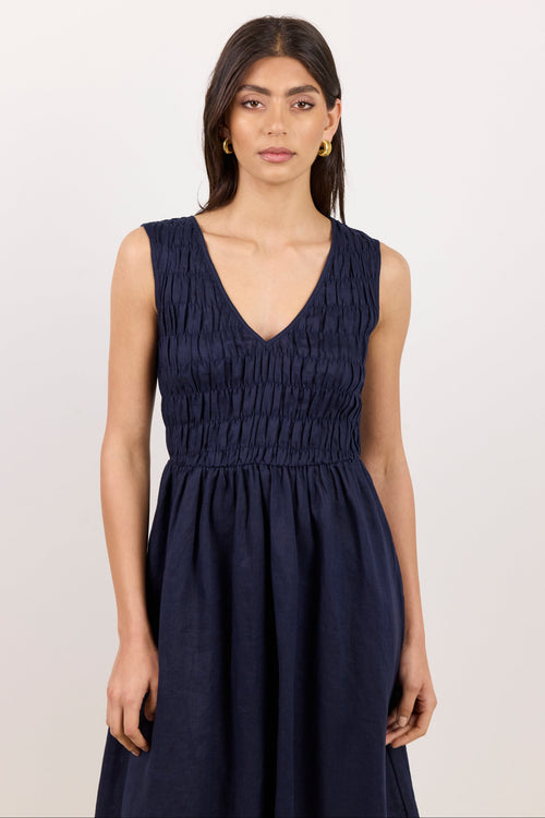 A woman modeling a sleeveless navy blue dress with a smocked bodice and V-neckline, photographed against a plain light background.