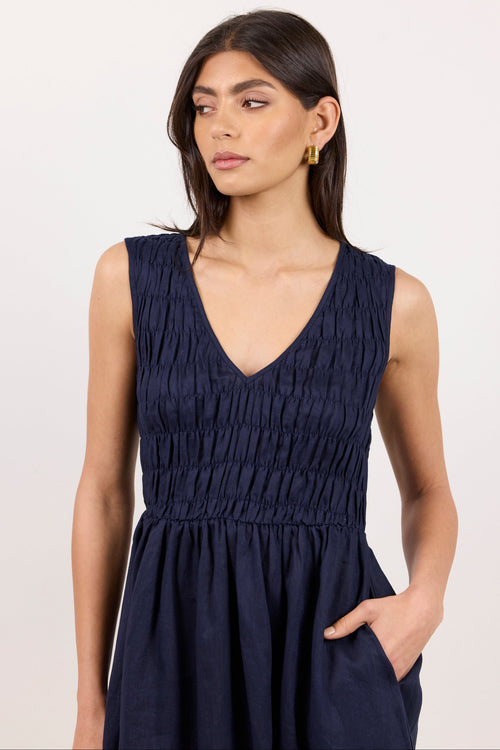 A woman wearing a sleeveless navy blue dress with a smocked bodice and V-neckline, standing against a plain light background.