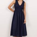 A woman modeling a sleeveless navy blue midi dress with a smocked bodice and V-neckline, paired with black sandals and sunglasses, standing against a plain white studio background.