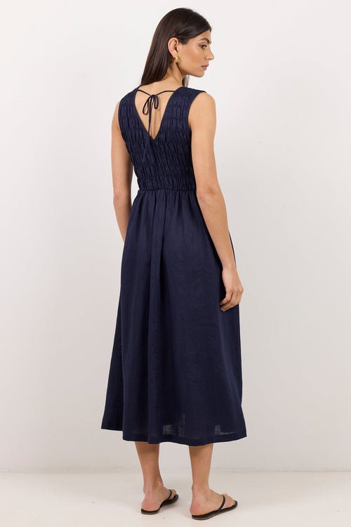 A woman modeling a sleeveless navy blue midi dress from the back, featuring a smocked bodice and tie-back detail, set against a plain off-white studio background.