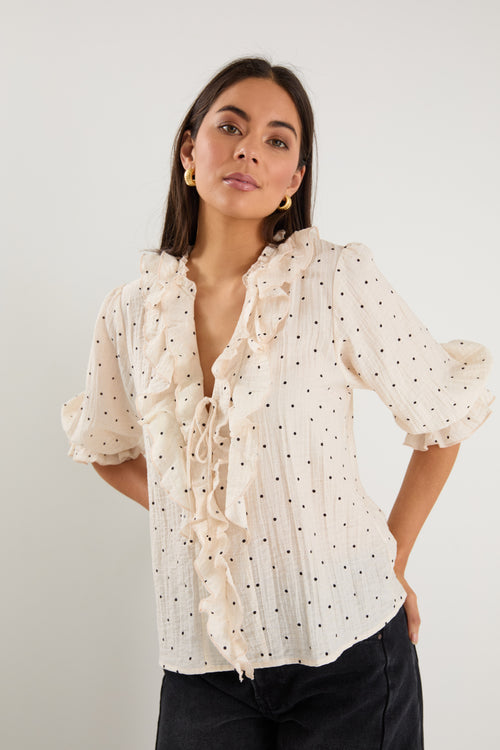 Woman wearing a cream blouse with black spot print, mid sleeves, button front, and ruffle trim with tie detail at the neckline.