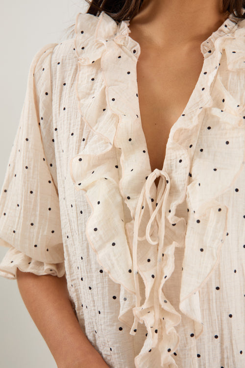 Woman wearing a cream blouse with black spot print, mid sleeves, button front, and ruffle trim with tie detail at the neckline.