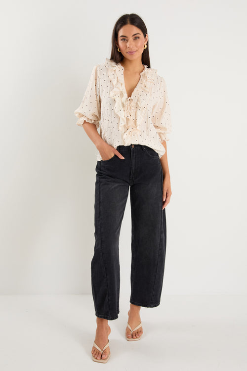 Woman wearing a cream blouse with black spot print, mid sleeves, button front, and ruffle trim with tie detail at the neckline.