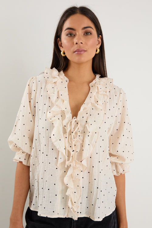 Woman wearing a cream blouse with black spot print, mid sleeves, button front, and ruffle trim with tie detail at the neckline.