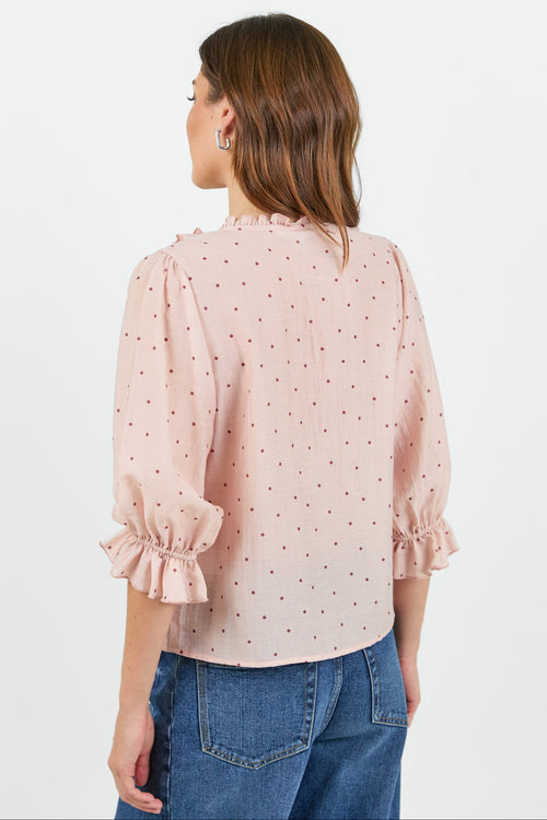 A woman shown from the back wearing a light pink blouse with small dark polka dots, ruffled trim at the collar and cuffs, paired with blue jeans against a plain white studio background.