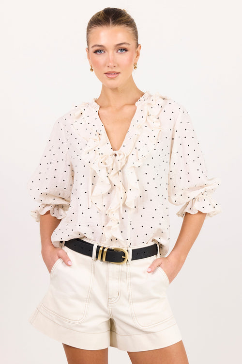 A woman modeling a white long-sleeve blouse with black polka dots and ruffle details, paired with white shorts and a black belt, against a plain white studio background.