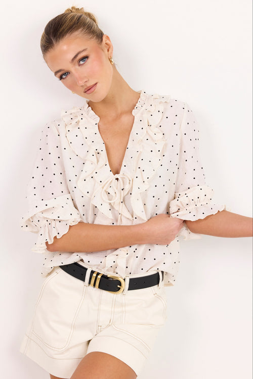 A woman modeling a white short-sleeve blouse with black polka dots, ruffled trim, and a tie neckline, paired with cream shorts and a black belt with a gold buckle, against a plain white background.