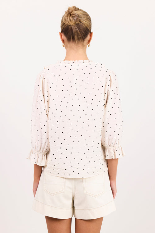 A woman viewed from behind wearing a cream-colored blouse with a small black polka dot pattern and cream shorts, set against a plain white studio background.