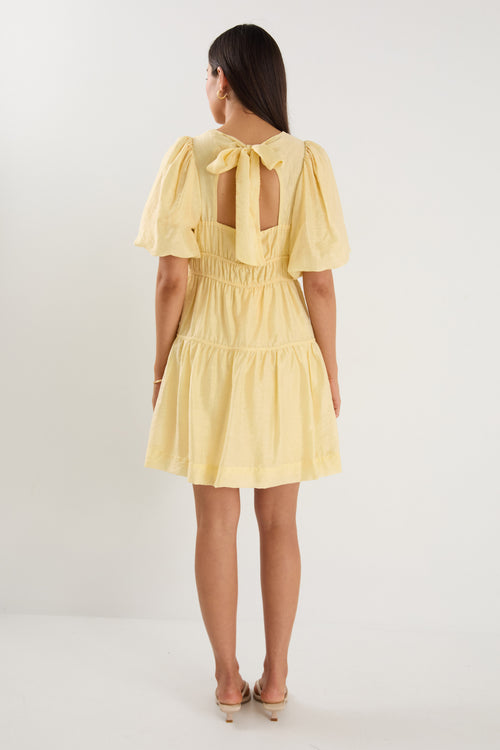 Woman wearing a short butter-yellow mini dress with puff sleeves, elastic waist, and back tie detail, styled with neutral sandals.