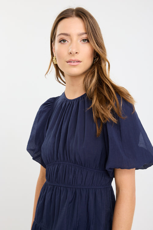 Woman wearing a navy mini dress with puff sleeves, elastic waist, and tie-back detail, styled with black flats.
