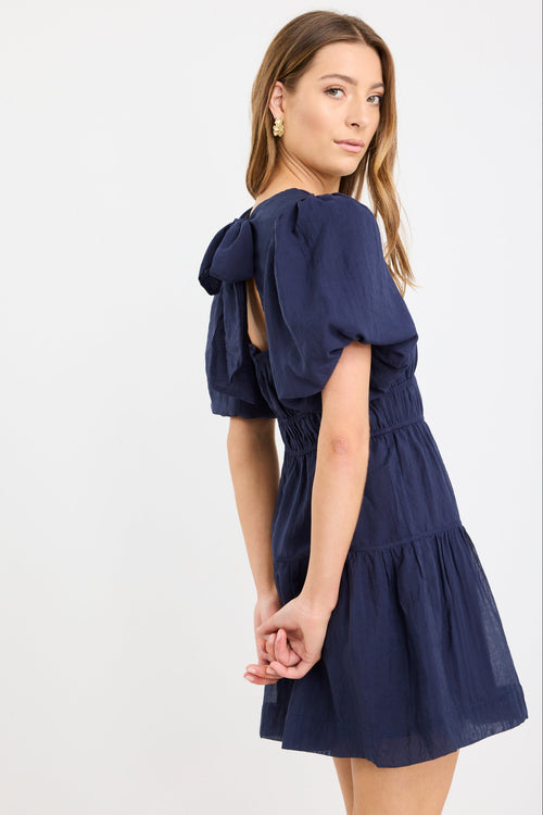 Woman wearing a navy mini dress with puff sleeves, elastic waist, and tie-back detail, styled with black flats.
