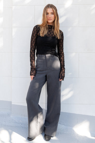 Woman wearing a black lace top and gray pants standing against a white wall.