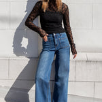 Woman wearing a black lace top and blue jeans standing against a light gray wall.