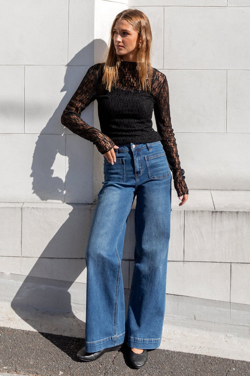 Woman wearing a black lace top and blue jeans standing against a light gray wall.
