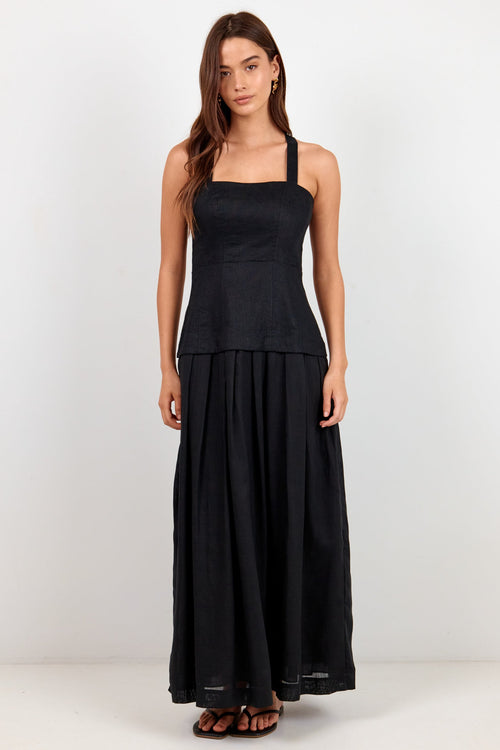 A woman modeling a black sleeveless maxi dress with a square neckline, fitted bodice, and flowing skirt, standing against a plain light gray studio background.