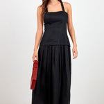 A woman modeling a black sleeveless maxi dress with a structured bodice and flowing skirt, styled with black sunglasses, brown sandals, and a red clutch bag against a plain white studio background.
