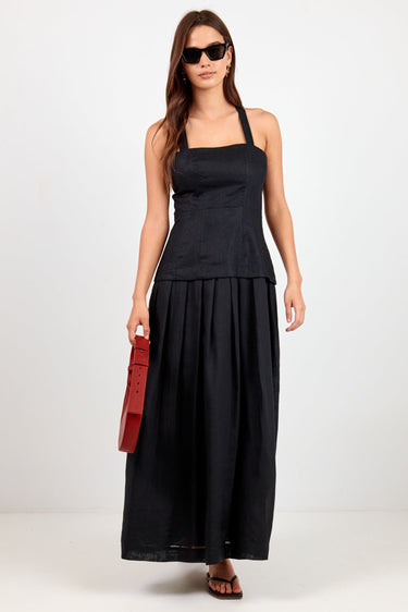 A woman modeling a black sleeveless maxi dress with a structured bodice and flowing skirt, styled with black sunglasses, brown sandals, and a red clutch bag against a plain white studio background.
