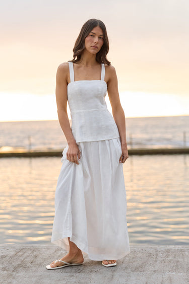 A woman wearing a white sleeveless linen top and matching white maxi skirt stands on a concrete pier with the ocean and sunset in the background.