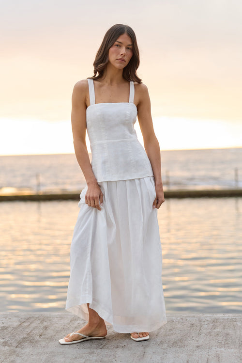 A woman wearing a white sleeveless linen top and matching white maxi skirt stands on a concrete pier with the ocean and sunset in the background.