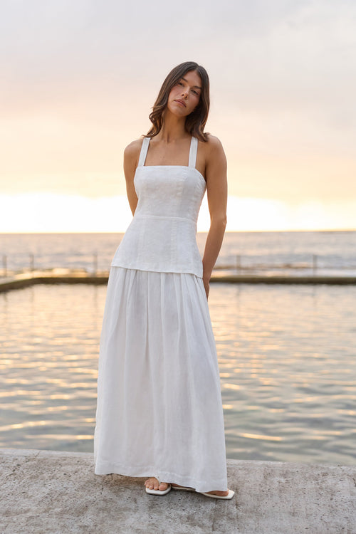 A woman wearing a white sleeveless maxi dress with a structured bodice and flowing skirt, standing outdoors by a calm body of water at sunset.